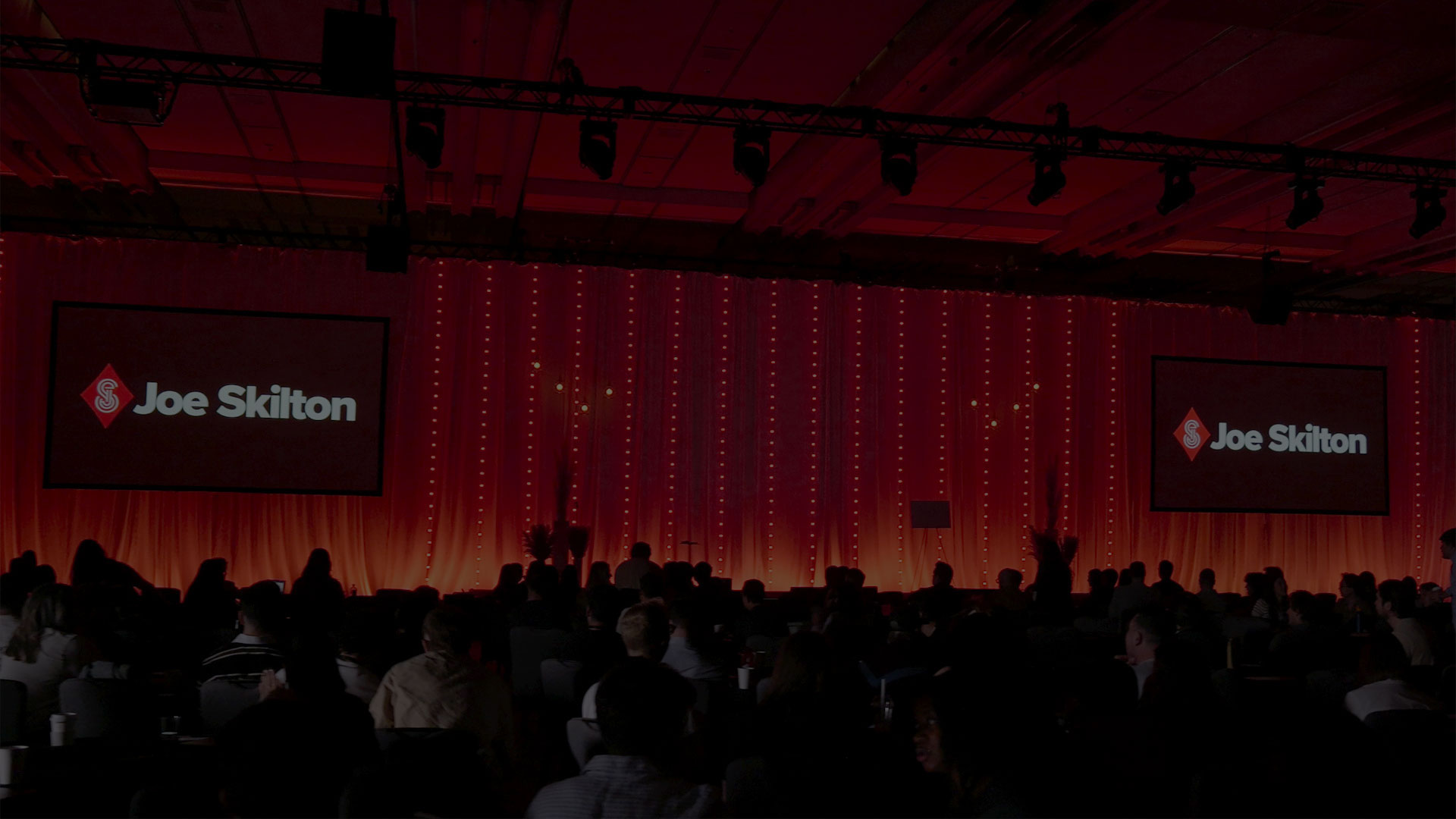 Corporate event stage in red wash lighting, with mentalist Joe Skilton's logo on IMAG screens.